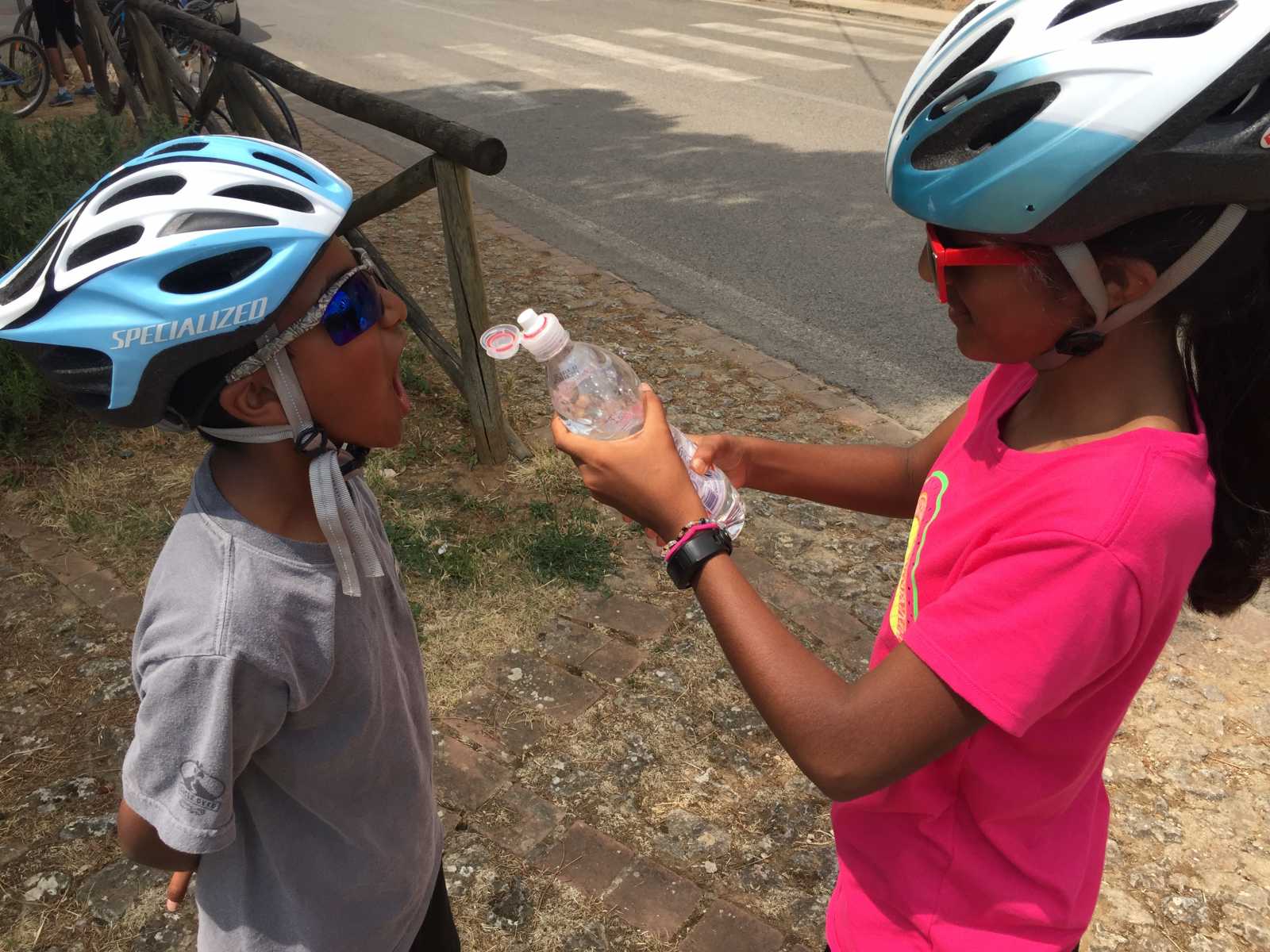 Two children in helmets are sharing water from a bottle on the roadside, taking a refreshing break during a "family biking tour" in Tuscany.