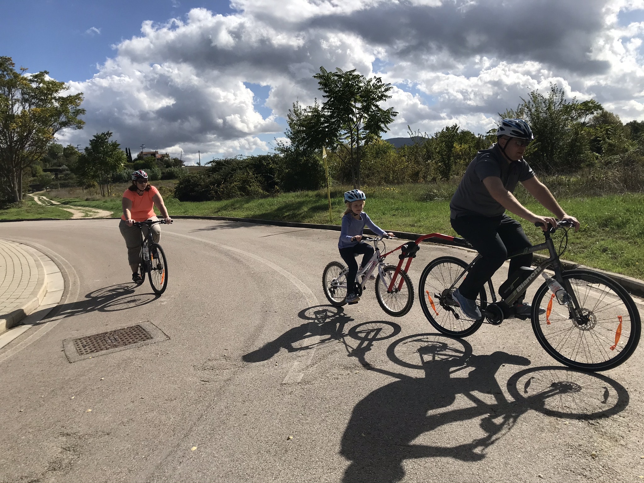 A family, including a child on a tag-along bike, rides along a paved road in the sunny Tuscan countryside, showcasing a great option for family biking tours.